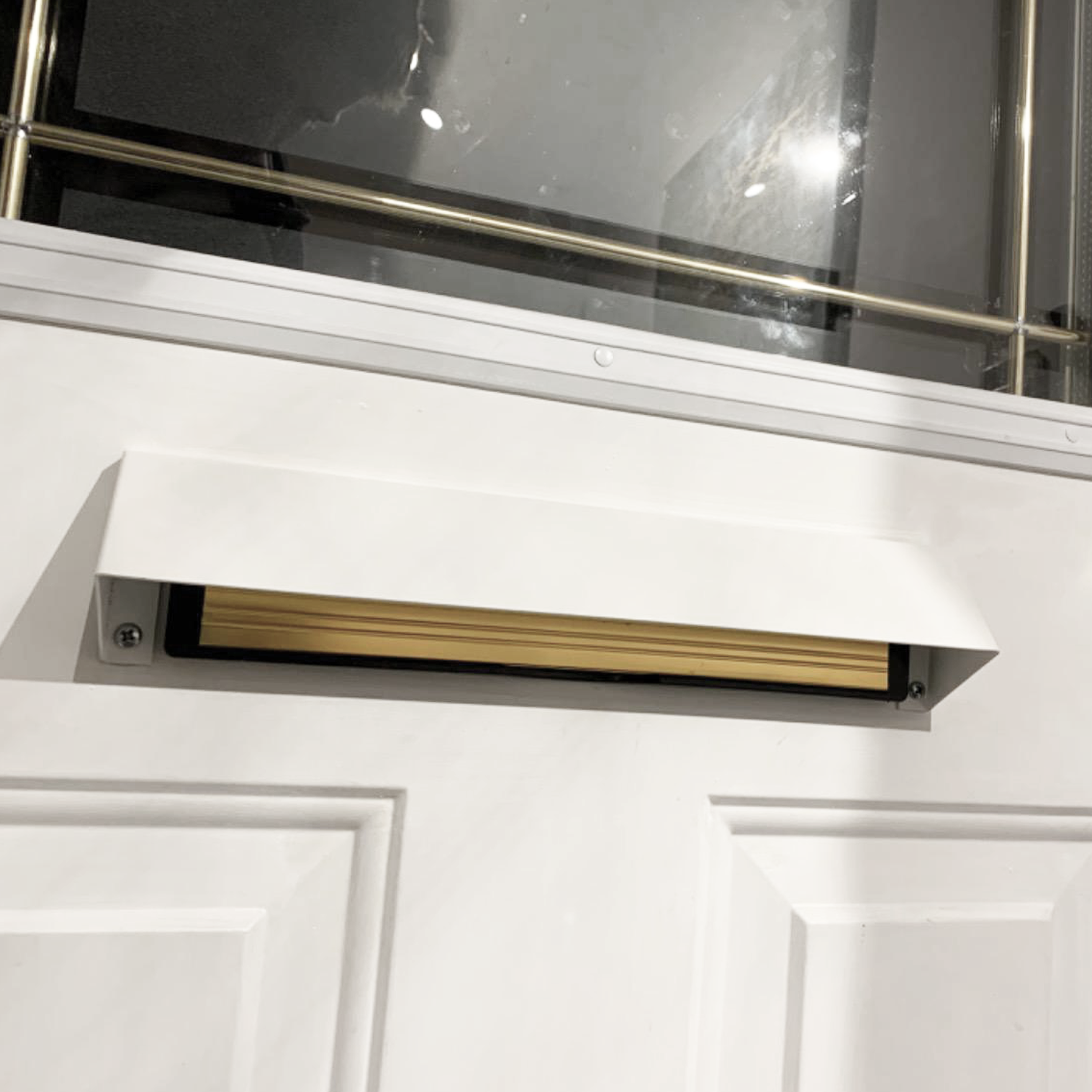 White front door with an open mail slot cover, revealing a wooden draft blocker or brush insert inside. The slot is positioned below a decorative glass window with metal framing