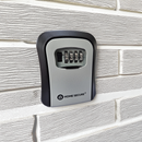 Wall-mounted Home Secure® outdoor key safe box attached to an exterior brick wall. The safe features a black and grey design with a 4-digit combination lock on the front