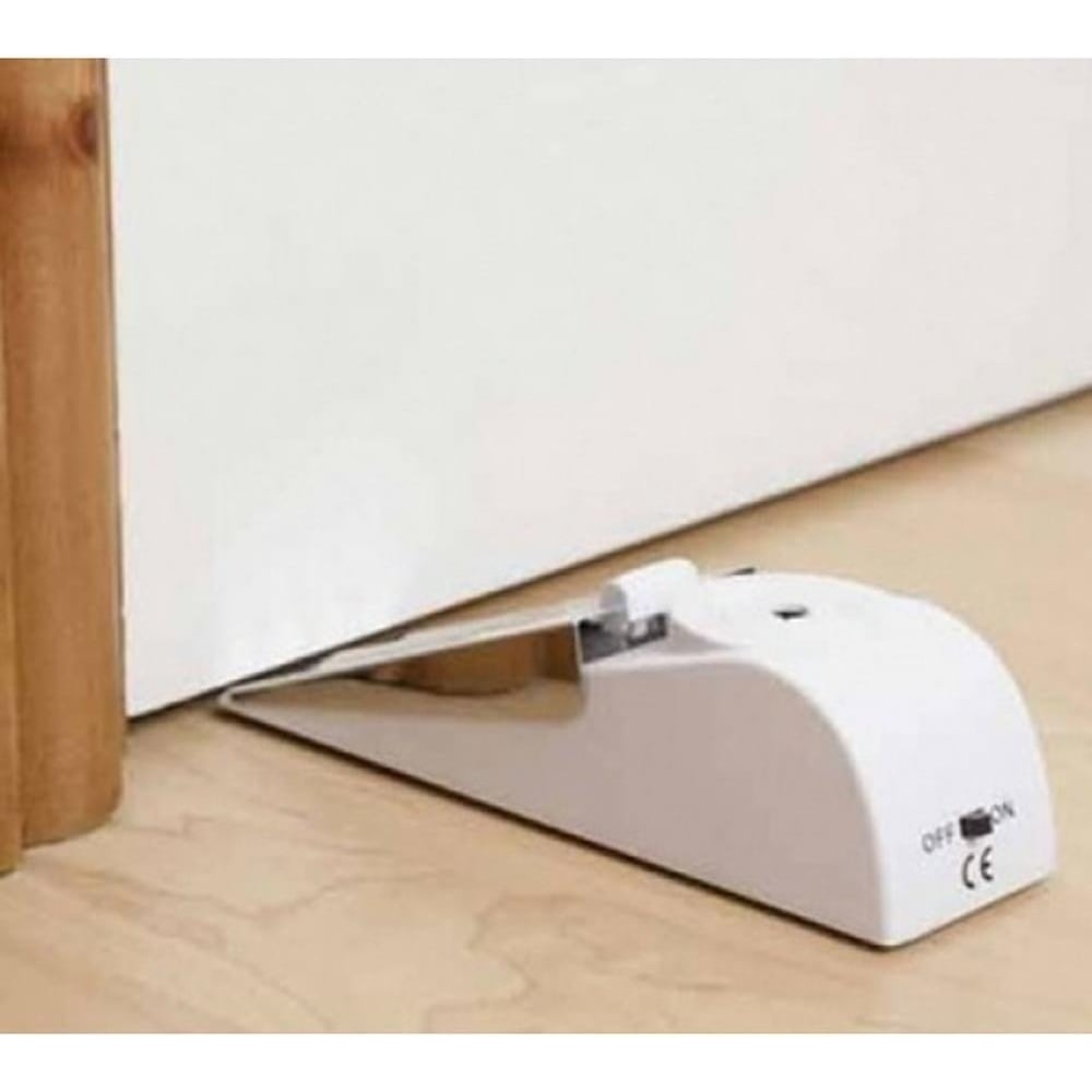 White door stop wedge alarm placed on a wooden floor, positioned under a white door. The device features an on/off switch and is designed to prevent the door from opening while sounding an alarm if tampered with.
