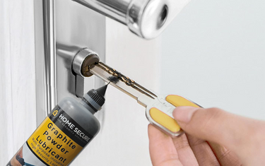 Hand inserting a key into a silver door lock while applying Home Secure graphite powder lubricant to the keyhole.