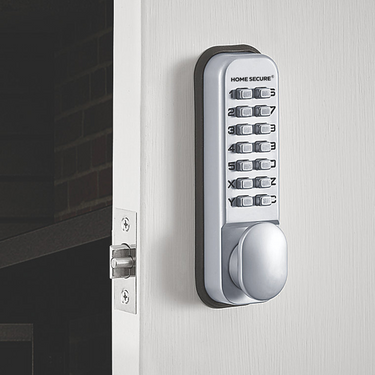 Digital mechanical door lock by Home Secure installed on a white door. The lock features a silver keypad with numbered push buttons (0–9) and a thumb turn at the bottom. The latch is visible on the door edge, aligned with the strike plate on the door frame